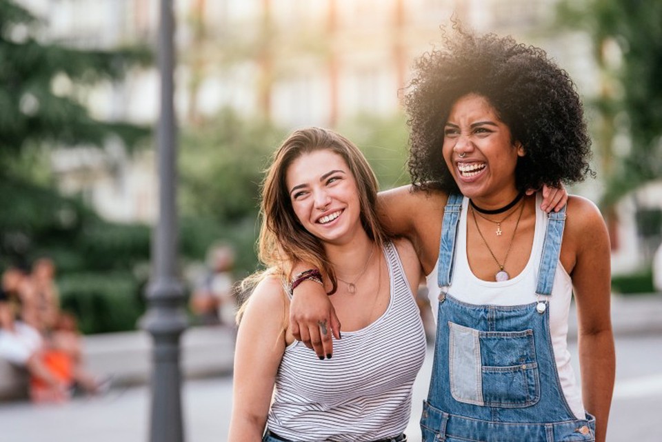 Same-sex couple smiles and embraces on sunny day in city.