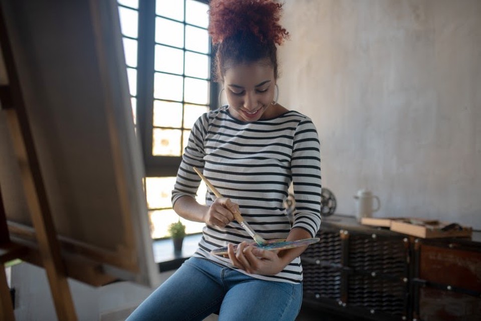 A person holds a paintbrush and palette while sitting in front of an easel.
