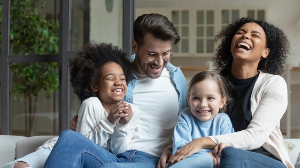 A family with two young daughters laughs together while sitting on a couch.