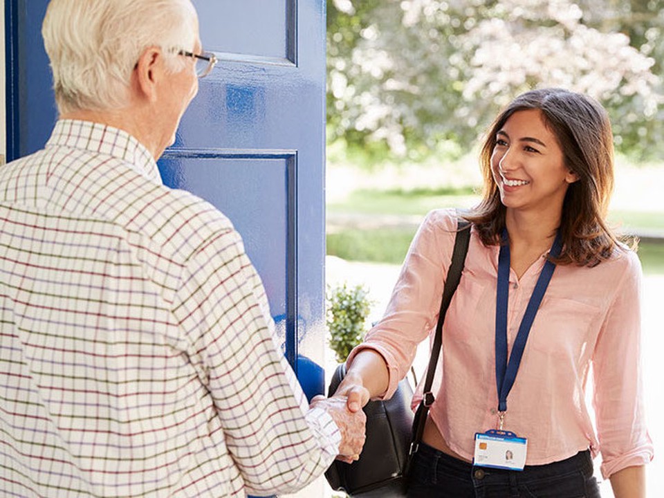 A female social worker greets an elderly man as she walks through his door.