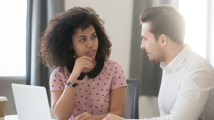 A corporate social worker sits next to an employee and listens to them speak at a desk near an open laptop.