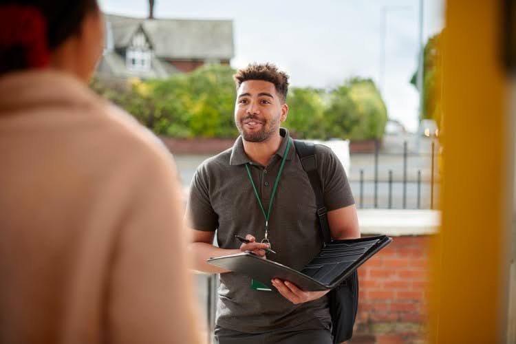A community social worker holding a clipboard talks with a resident in a doorway.
