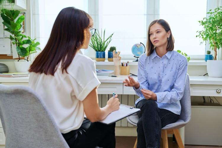 A social worker holding a clipboard talks to a client seated on a chair.
