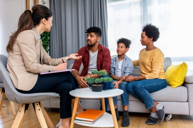 A social worker with a clipboard talks to two parents and a child seated on a couch.