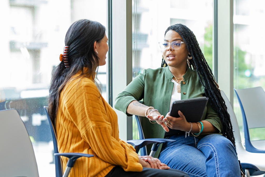 Two students converse in a public space; one is holding a tablet.